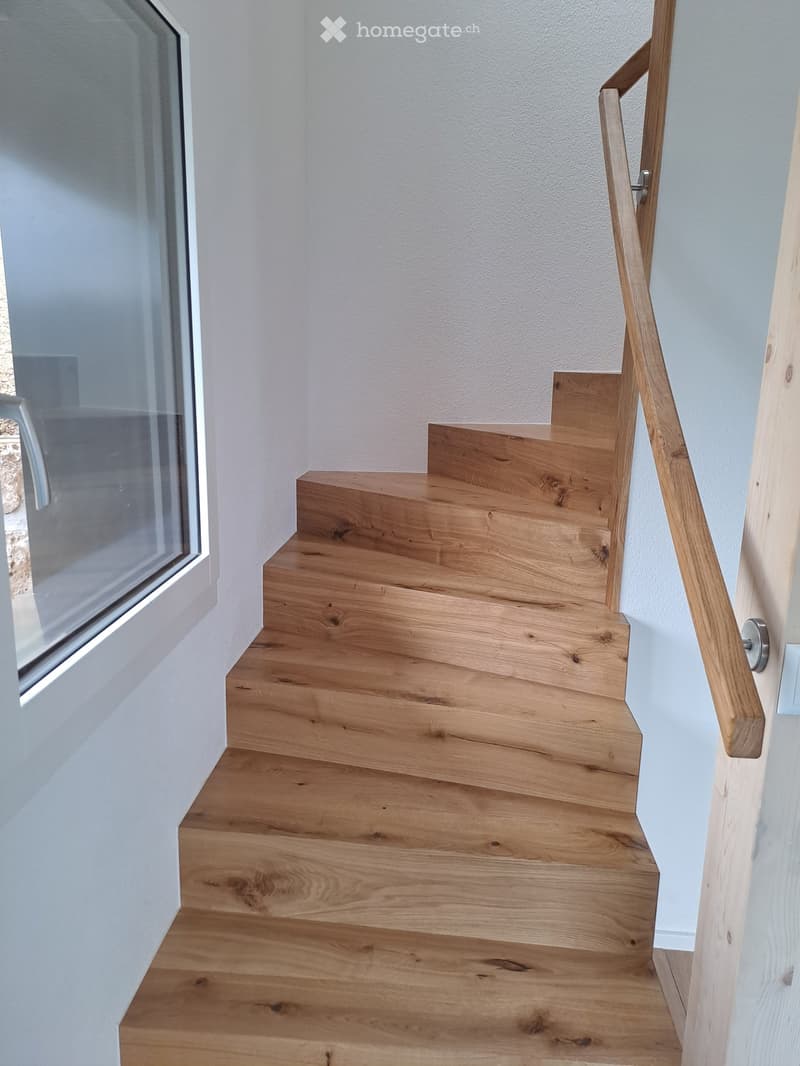Wooden staircase with a modern wooden handrail, light-colored walls, and a large window on the left side.