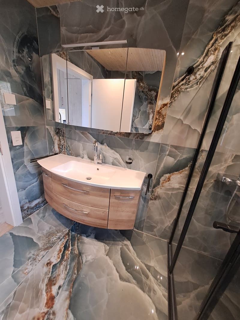 Modern bathroom with a white sink, wooden cabinet, and a large mirror above it. The walls and floor feature a marble-like pattern in shades of grey and brown.