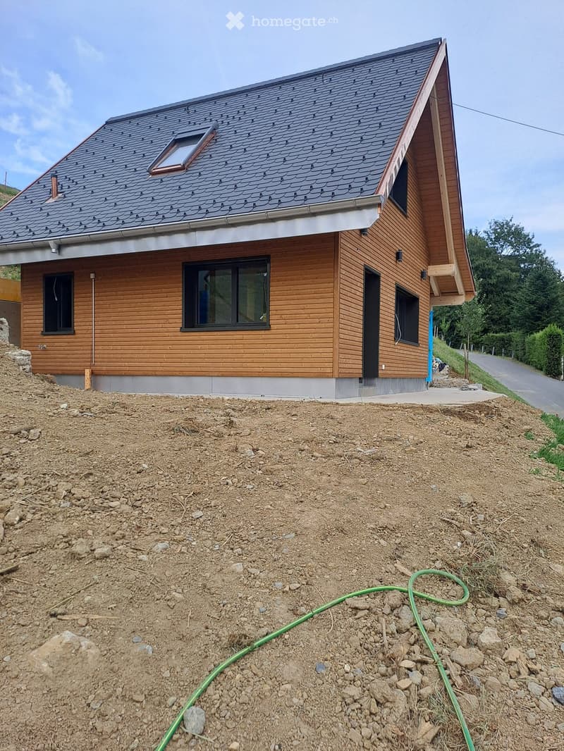 Modern two-story house with wooden siding, dark gray shingled roof, multiple windows, and a raised ground floor.