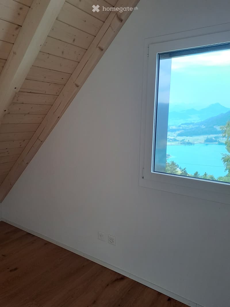 Empty attic room with wooden slanted ceiling, light wooden floor, and large window offering a scenic view of a lake and mountains.