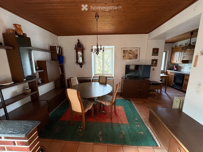 central dining table, wooden chairs, tiled floor, kitchen area, wall-mounted television, shelves with decorative items, cabinets, picture frames
