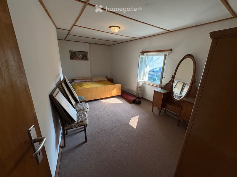 Single bed, bedsheets, wooden bed frame, picture frame on wall, mirror, small wooden cabinet, small rug, wooden chair, carpet on floor, window with curtains