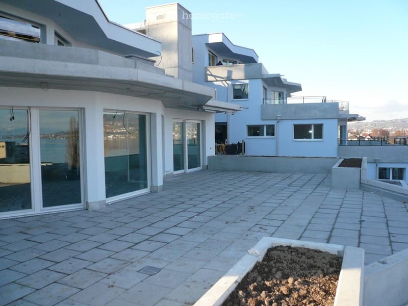 Modern outdoor patio area with tiled flooring, low concrete walls, and a small garden bed with soil. The patio is adjacent to a contemporary white building with large glass windows and balconies.