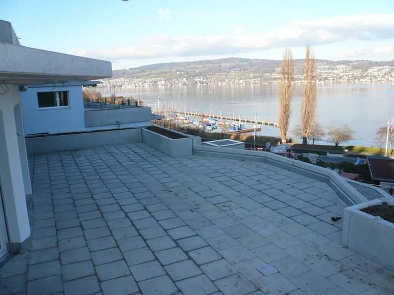 Terraced balcony with stone tiles, overlooking a waterfront with boats and distant hills.