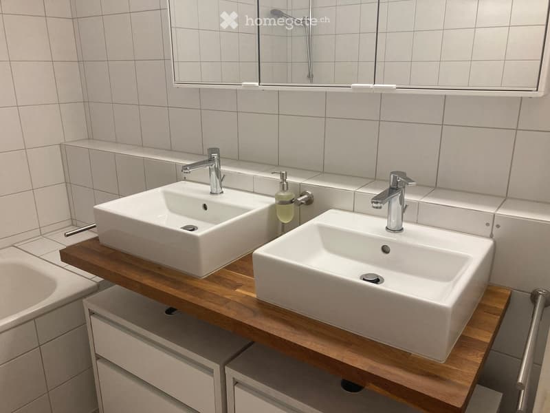 Modern bathroom with two white sinks on a wooden vanity, chrome faucets, a mirror, and a soap dispenser.