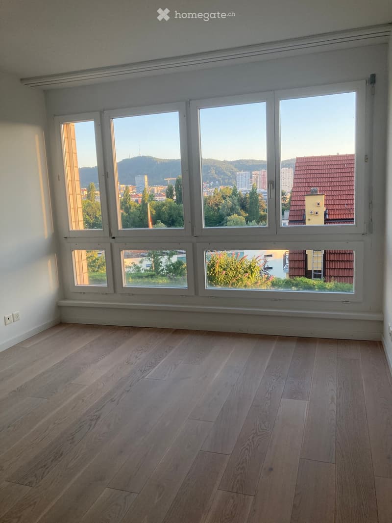 Empty room with wooden flooring and large windows offering a view of the city and surrounding landscape.