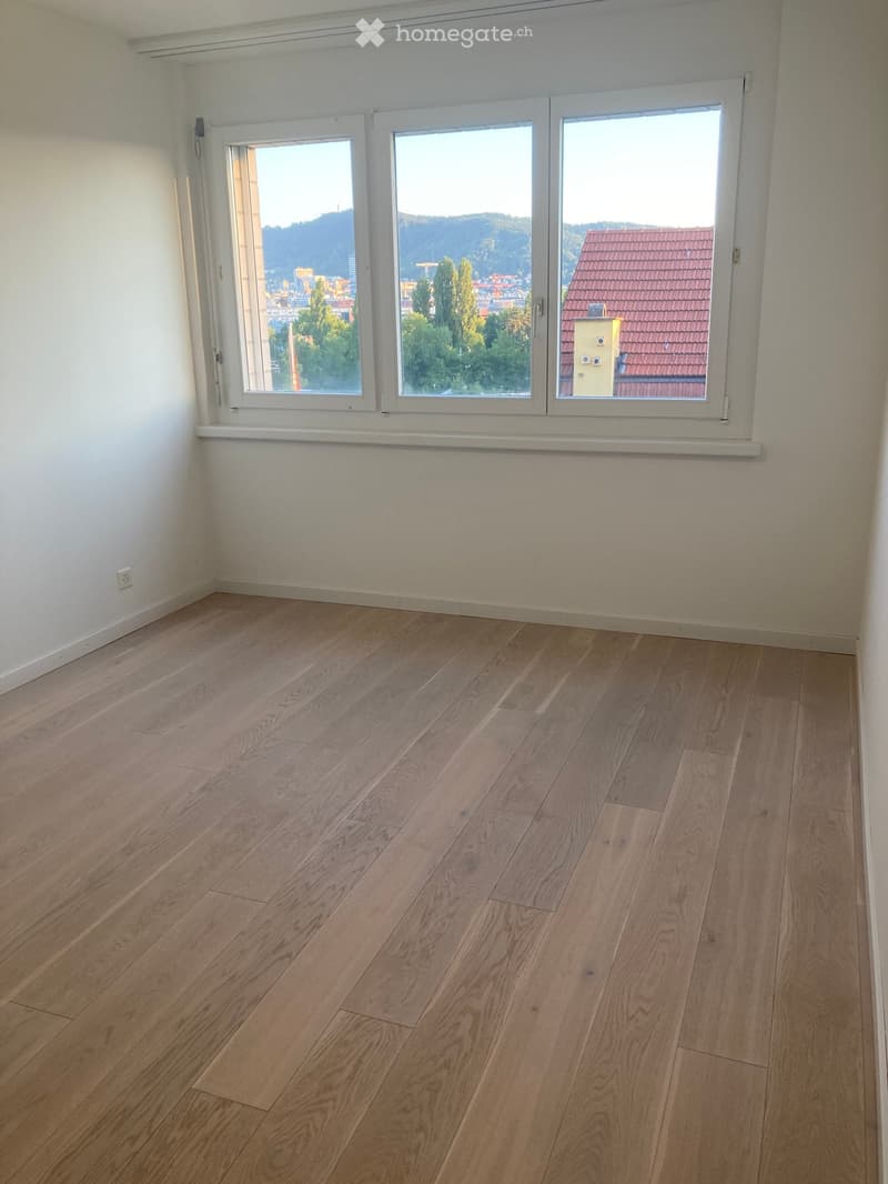 Empty room with light wooden flooring and large windows offering a view of the city and surrounding landscape.