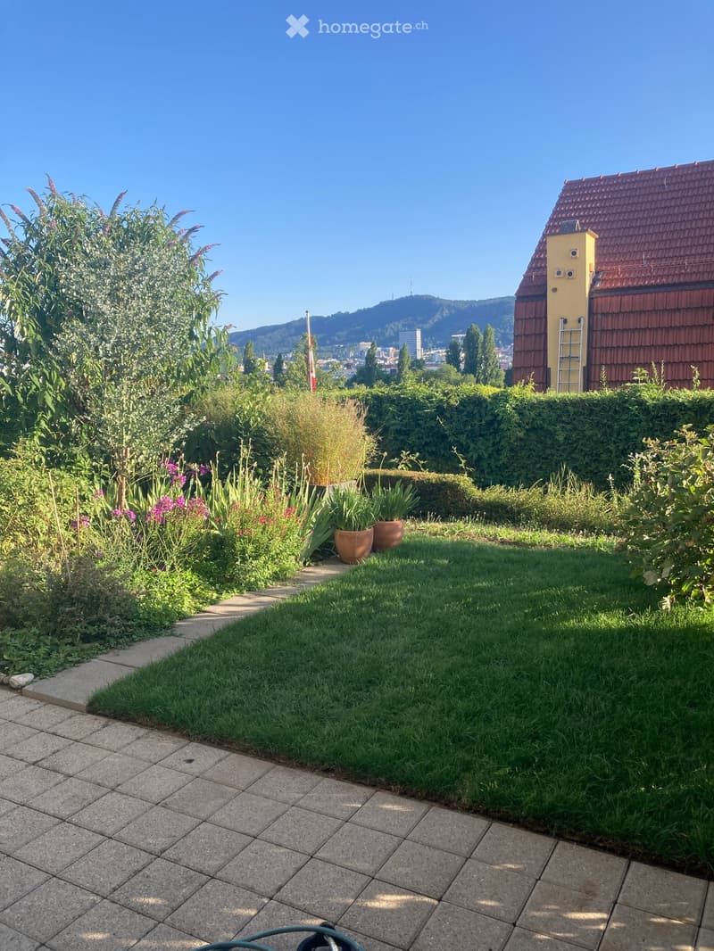 Well-maintained garden with a lawn, flower beds, potted plants, and a pathway leading to a house with a red-tiled roof.