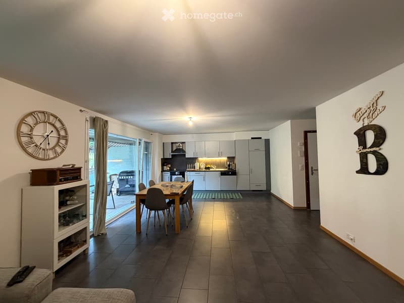 Open-plan living and dining area with a dining table and chairs, kitchen with cabinets and appliances, and a wine rack. Large windows lead to a patio with outdoor furniture.