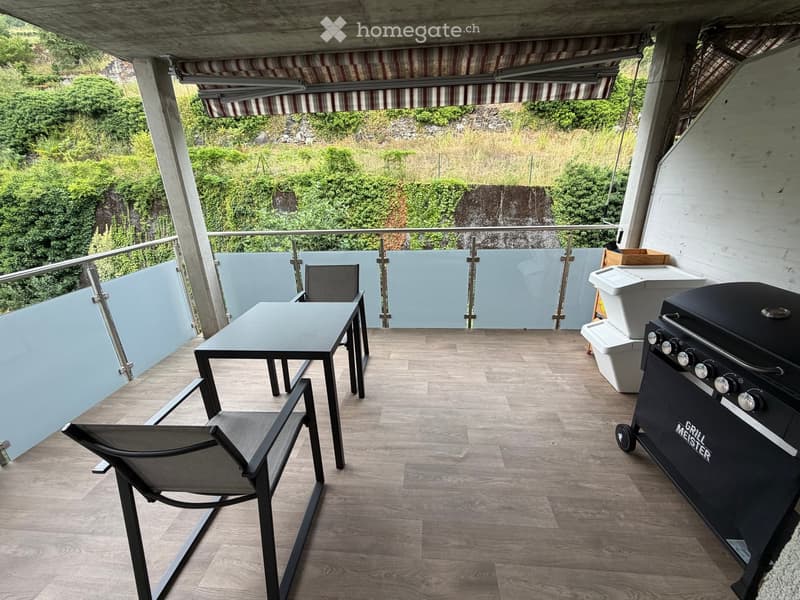 Balcony with wooden flooring, a table, two chairs, and a grill, featuring a view of greenery and a waterfall.