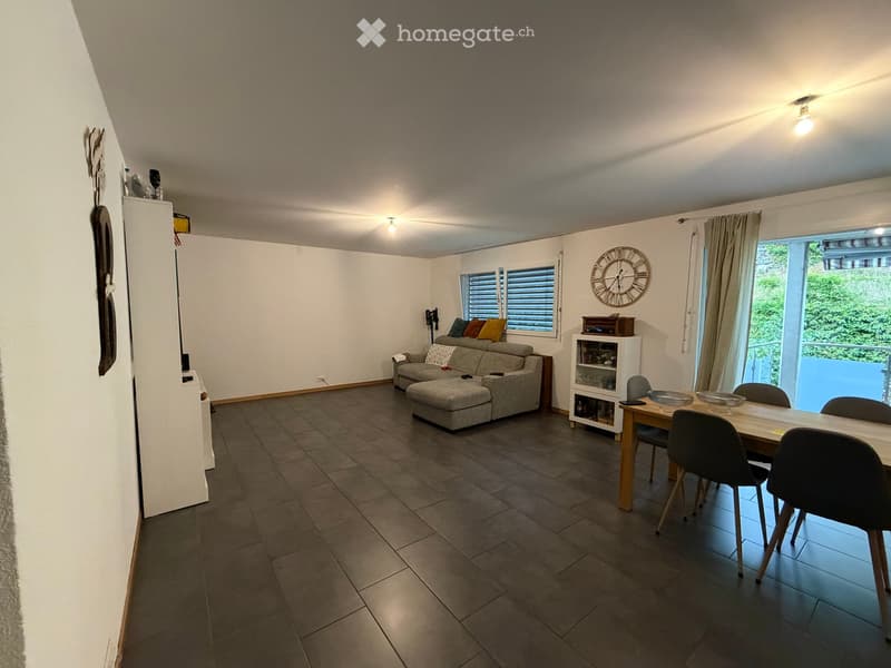 Open-plan living and dining area with a grey sofa, ottoman, dining table with chairs, and a shelf with glassware. The space features large windows and a modern aesthetic.