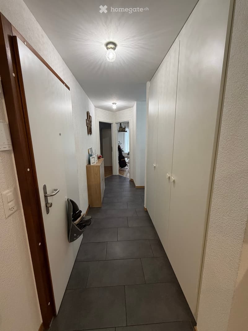 A narrow hallway with grey tiled flooring, white walls, and built-in white cabinets on one side. A small wooden cabinet with framed pictures and a shoe rack is visible on the left, and a door leads to another room at the end.