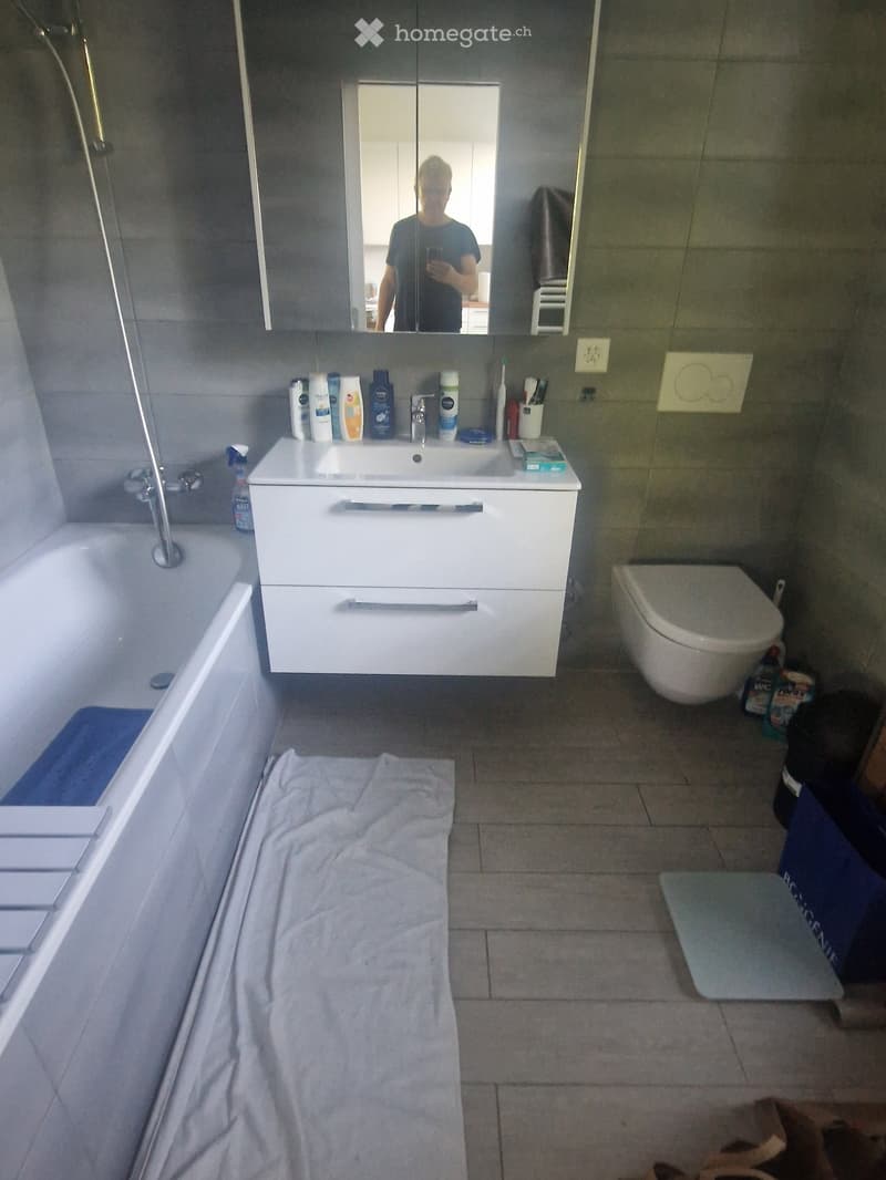 Modern bathroom with a white sink, mirror, toilet, bathtub, and various toiletries on the sink counter. A towel is placed on the floor.