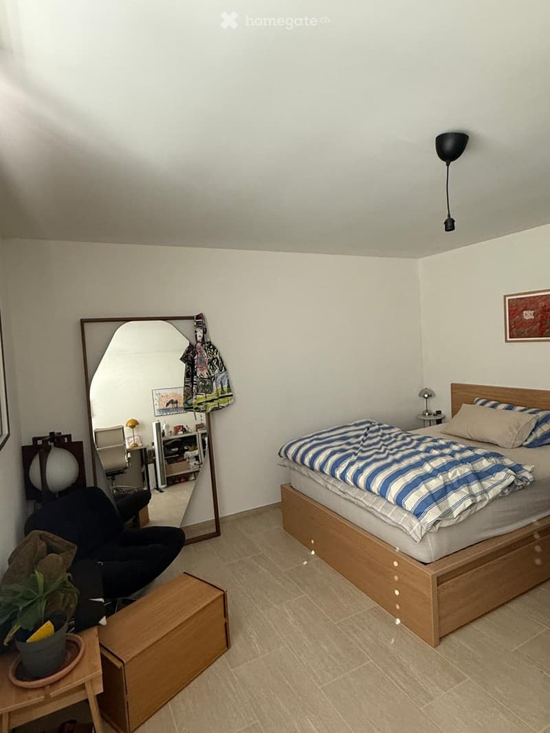Cozy bedroom with a wooden bed, striped bedding, a large mirror, a small table with a plant, and a chair. The room has light-colored walls and tiled flooring.