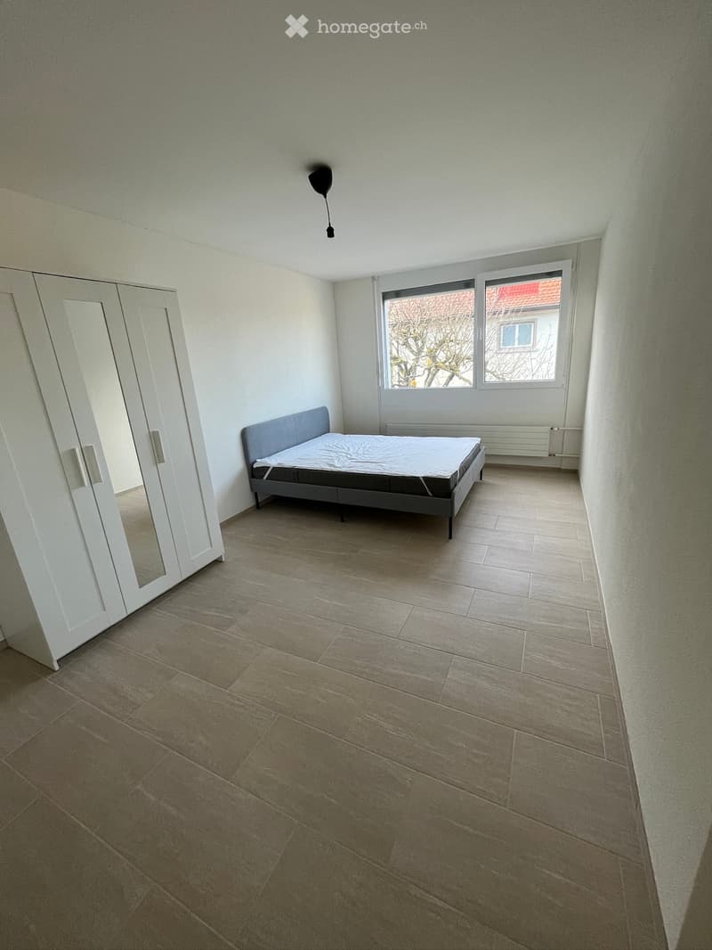 A furnished bedroom with a bed, wardrobe, tiled floor, and large windows.