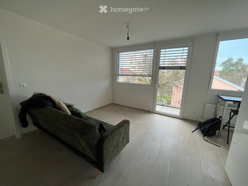 Living room with a sofa, a small table, a chair, and a backpack, featuring large windows and a sliding glass door leading to a balcony.