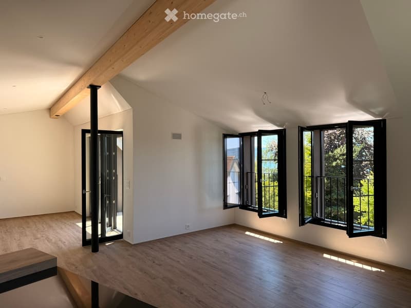 Bright, empty room with wooden flooring, white walls, large windows, and a black metal column. No furniture or equipment present.