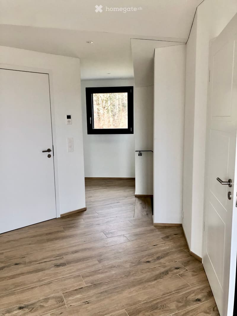 Empty hallway with light wooden flooring, white walls, and a large window at the end. Doors on both sides lead to other rooms.
