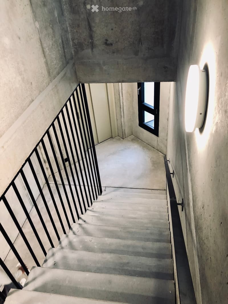 Concrete staircase with black metal railing, small window at the top, and a wall-mounted light fixture on the right side.