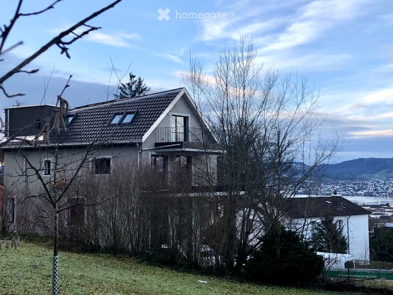 Two-story house with a modern design, featuring a sloped roof, multiple windows, and a balcony on the upper level. The house is surrounded by trees and offers a scenic view of the city and water in the background.