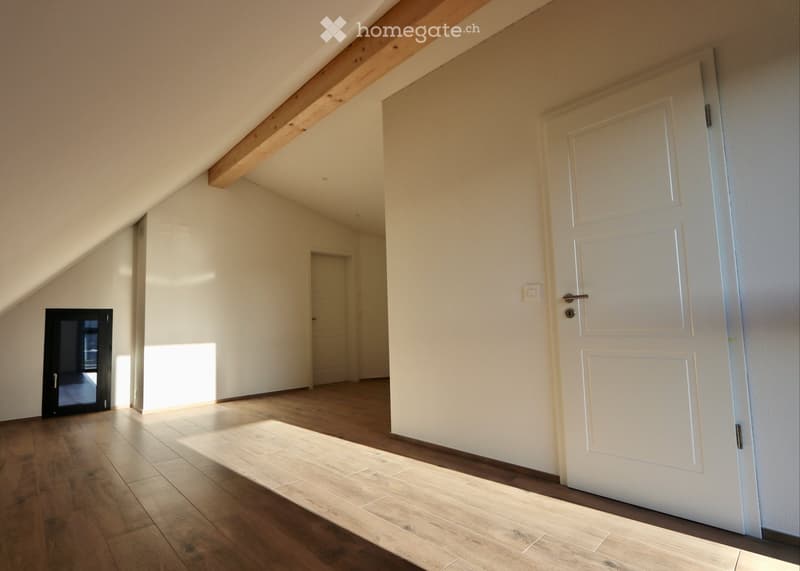 Empty hallway with wooden flooring, white walls, and exposed wooden beams on the ceiling. Features a white door and a black-framed window letting in natural light.