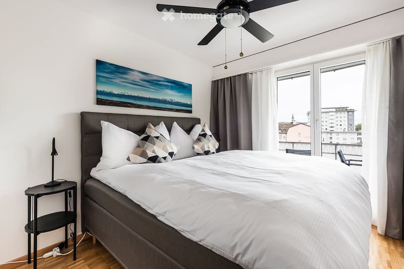 Modern bedroom with a large bed, white bedding, decorative pillows, nightstand with lamp, and a scenic painting above the bed. Large windows with curtains offer a view of the city.