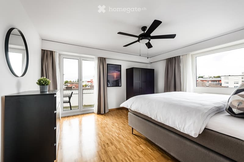 Modern bedroom with a large bed, wooden floor, large windows, and a balcony door leading to a small outdoor area. Features include a wardrobe, a round mirror, a small table with a potted plant, and a ceiling fan.