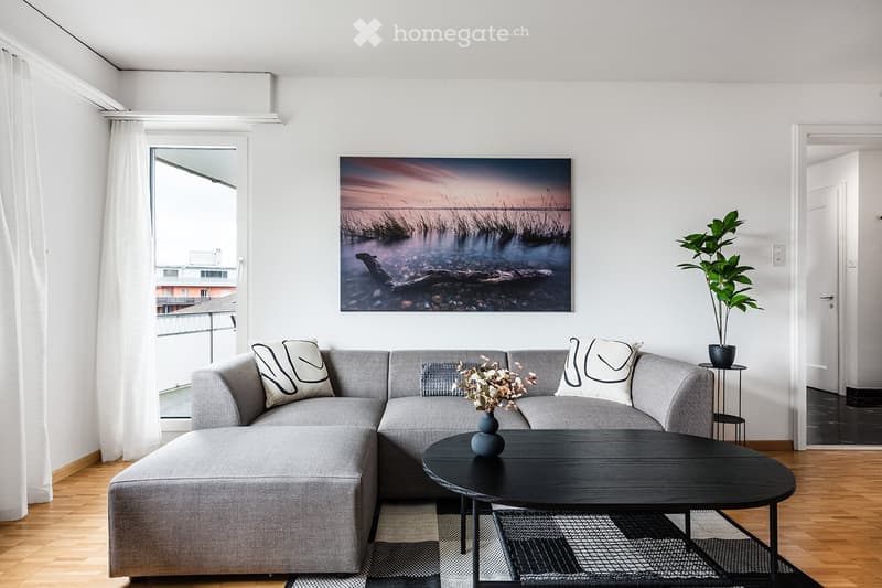 Modern living room with grey sectional sofa, black coffee table, decorative vase with flowers, potted plant, and artwork on the wall. Large window with white curtains offers a view of the balcony and cityscape.