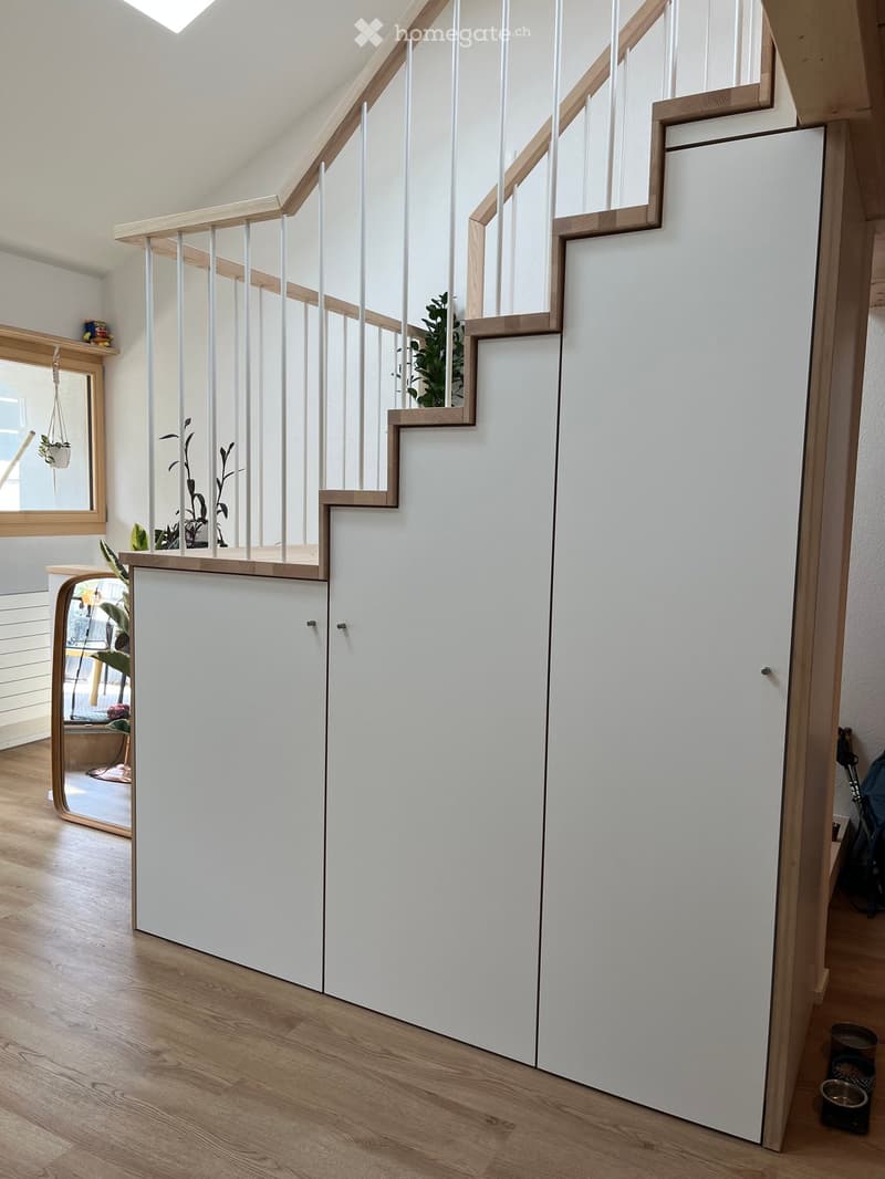 Modern hallway with a staircase featuring white railings and wooden steps, white walls, and a wooden floor. There are small potted plants and a mirror visible in the background.