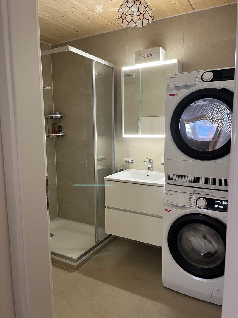 Modern bathroom with a glass-enclosed shower, a white sink with a modern faucet, and a stacked washer/dryer unit. The space features light-colored tiles and a sleek, contemporary design.