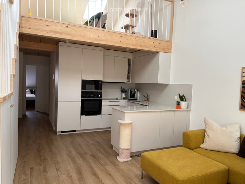 Modern, open-plan living and kitchen area with a yellow sofa, cat tree, sleek white kitchen cabinets, and a small island. The space features wooden flooring and large windows, with a lofted bedroom area visible in the background.