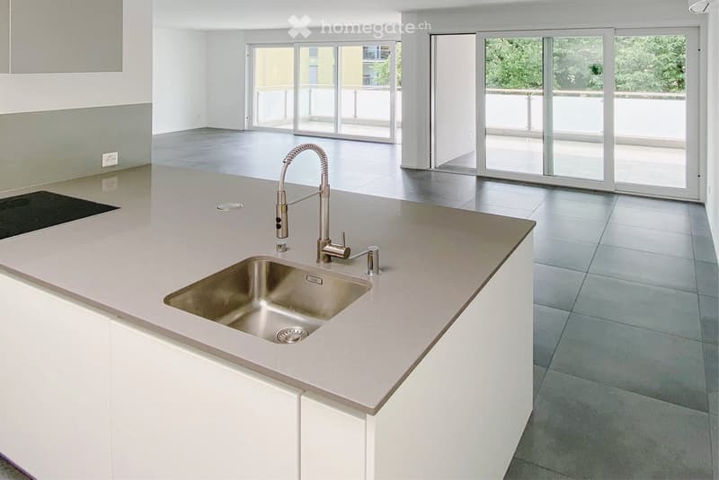 Modern kitchen with white cabinets, light grey countertops, and a stainless steel sink with a pull-out faucet. Large windows and sliding glass doors lead to a balcony.
