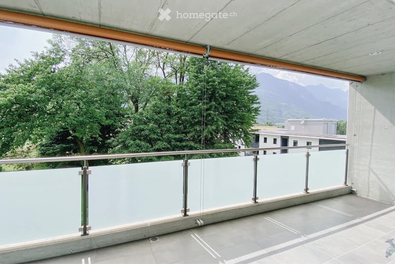 Balcony with glass railing, overlooking a green area and mountains.