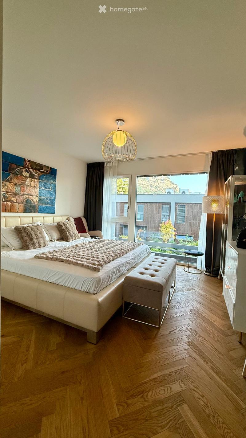 Modern bedroom with a large bed, bench, and stool, featuring wooden flooring and large windows with a view.