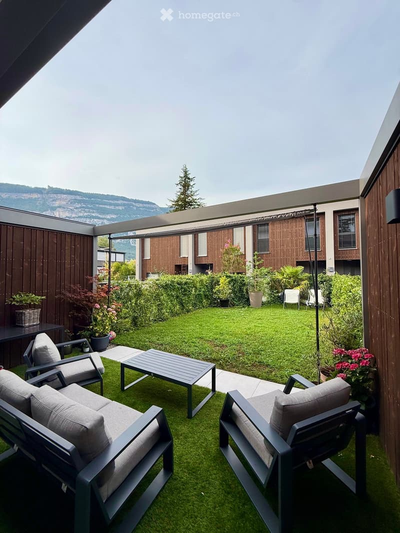A modern outdoor patio with artificial grass, grey furniture including a sofa, chairs, and a coffee table, surrounded by potted plants and a view of a landscaped garden and mountains.