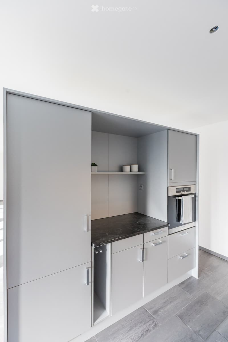 Modern kitchen with light grey cabinets, dark countertop, and built-in oven. Shelves hold small decorative items.