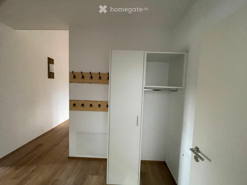 Empty hallway with wooden flooring, white walls, coat rack, and a white closet with mirrored door.