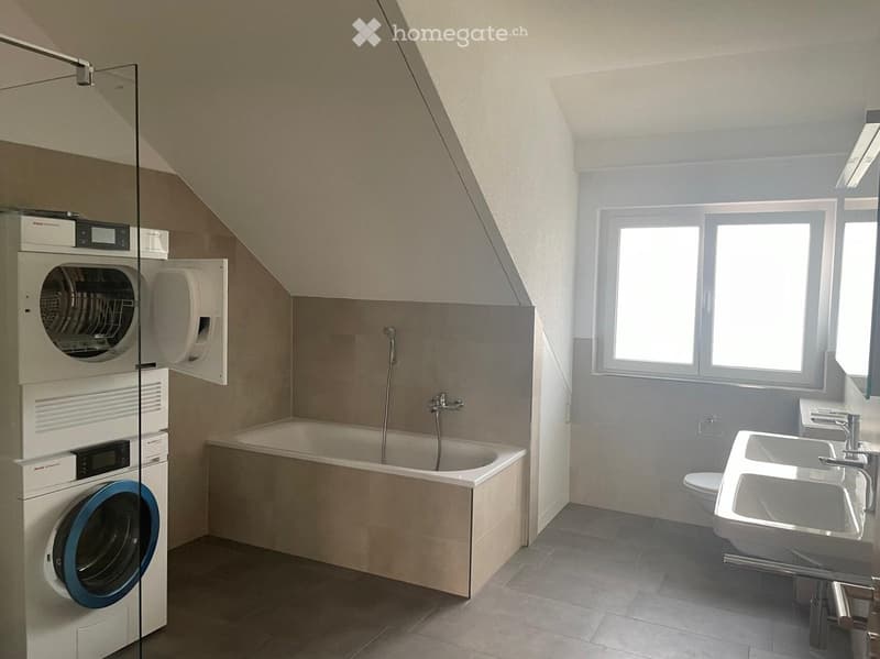 Modern bathroom with a bathtub, sink, toilet, and stacked washing machine and dryer unit. Large windows provide natural light.