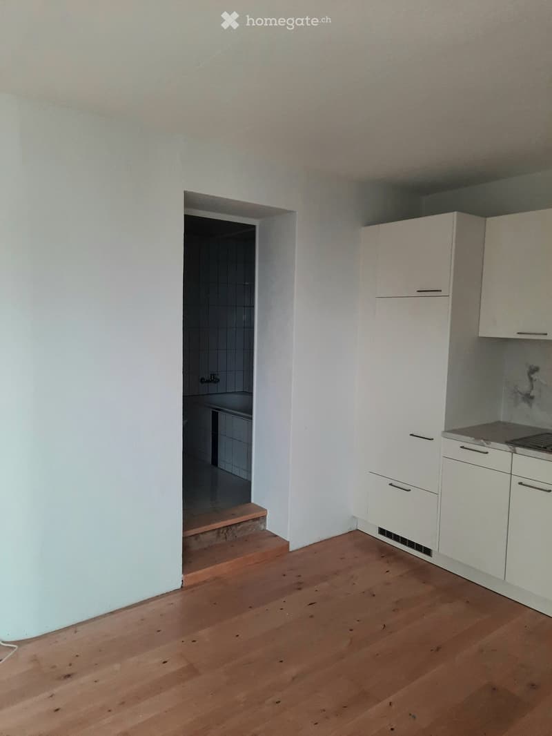 Empty room with wooden flooring, white walls, and white kitchen cabinets. A small staircase leads to a tiled bathroom area.