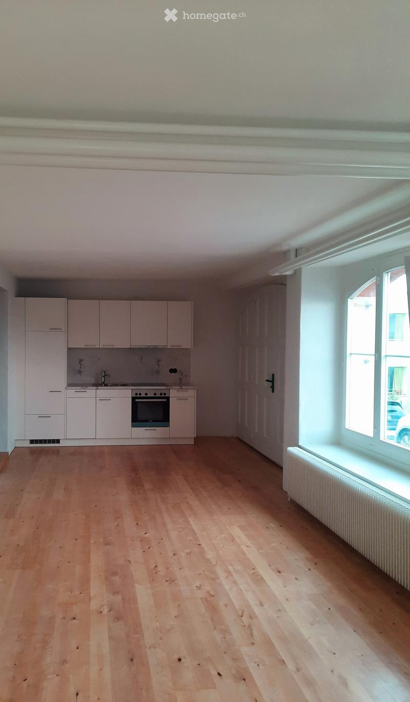 Empty kitchen with light wooden flooring, white cabinets, and a built-in oven. Large windows allow natural light to fill the space.