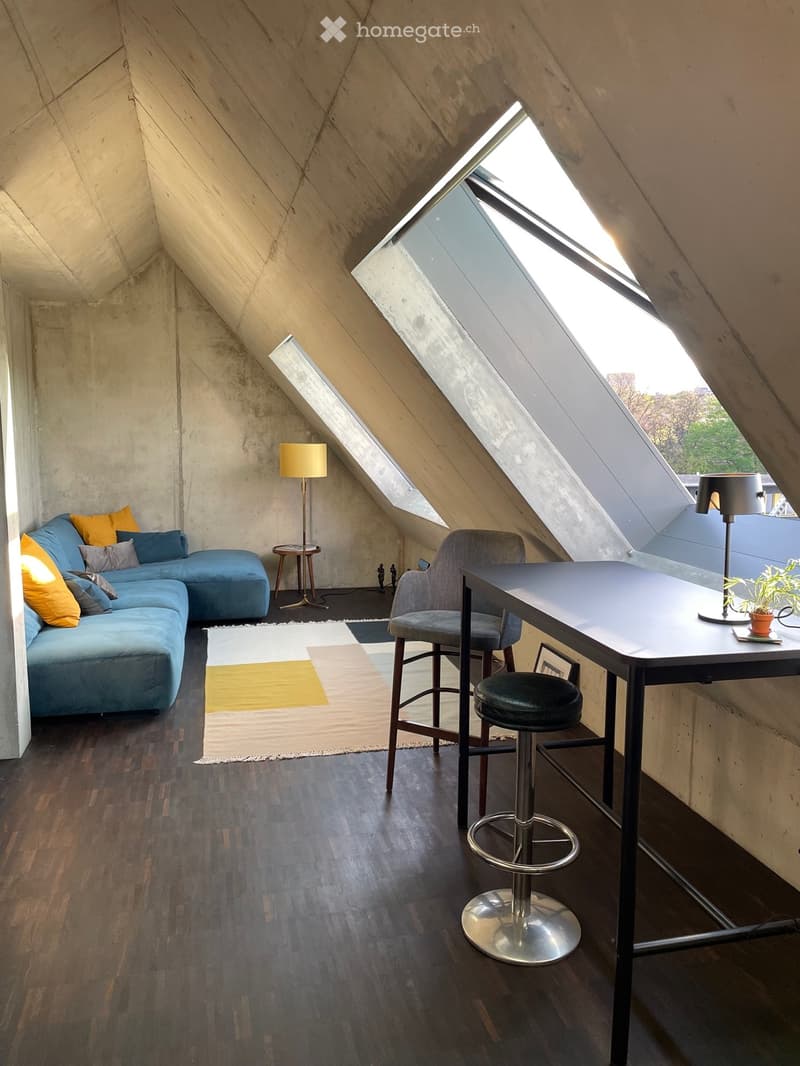 Loft-style room with wooden flooring, exposed concrete walls, skylights, a blue sectional sofa, a small round table, a desk with a chair, and a floor lamp.