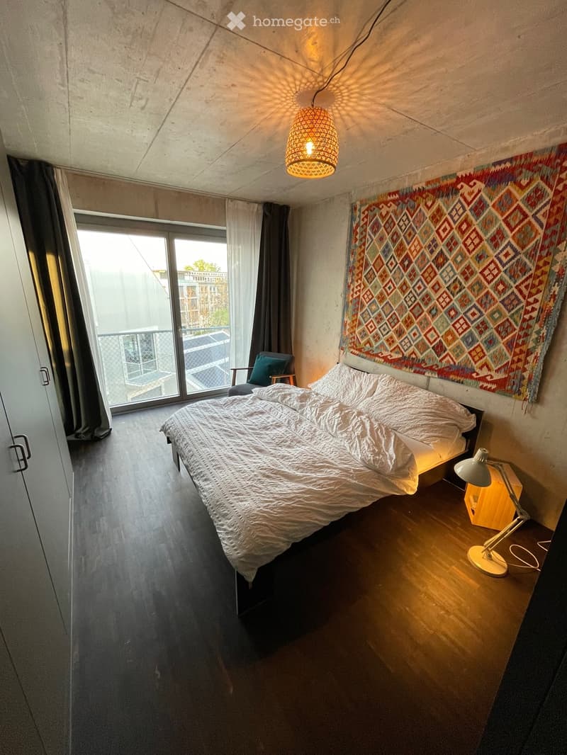 Modern bedroom with a double bed, nightstand with lamp, chair, large window with balcony access, and colorful wall tapestry.
