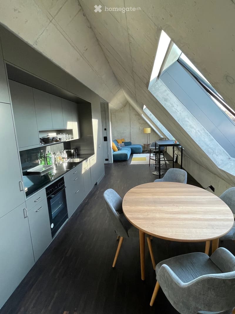 Modern, compact living space with a kitchen area, dining table, and seating area. Features grey cabinets, a stove, sink, and a small sofa with pillows.