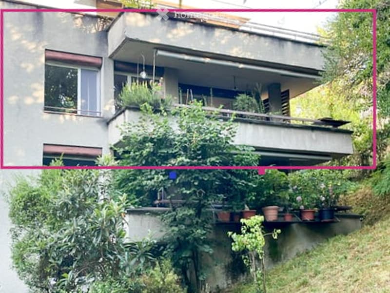 Multi-story residential building with balconies and greenery, featuring multiple windows and a raised ground floor.