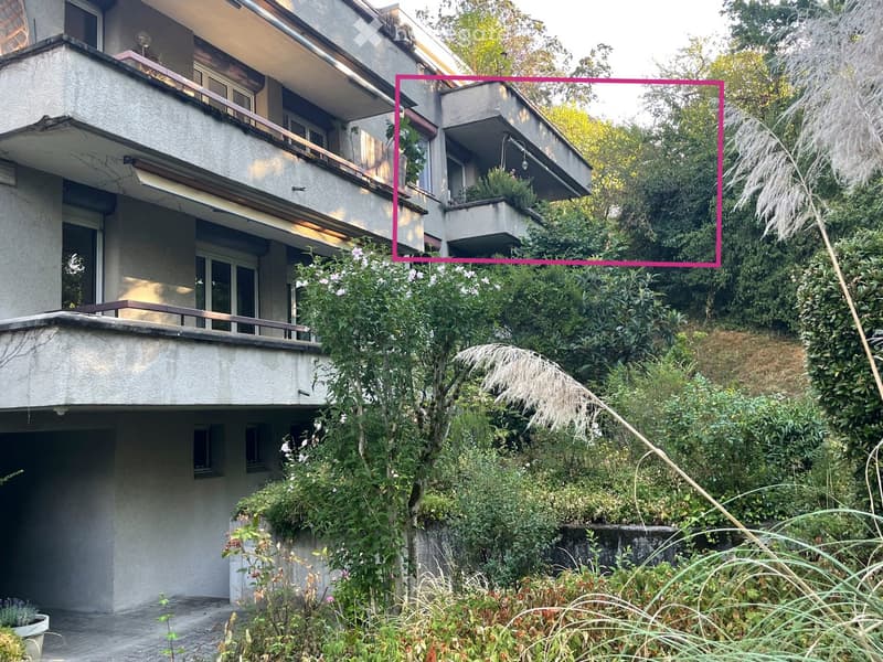 A multi-story residential building with balconies, surrounded by lush greenery and plants.