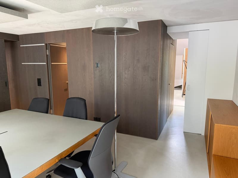 Modern office space with a large conference table, several ergonomic chairs, a floor lamp, and wooden cabinetry.