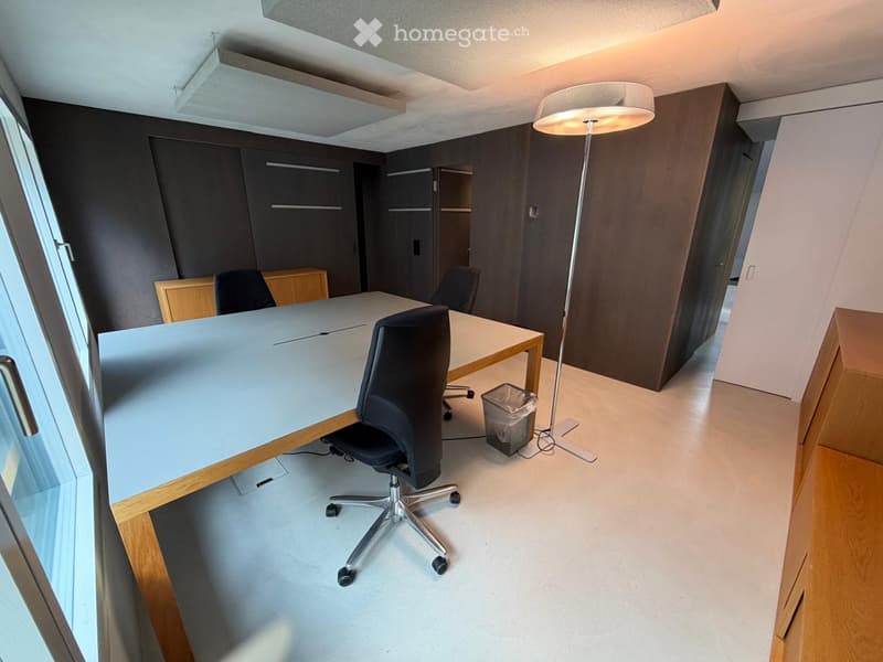 Modern office space with a large table, chairs, floor lamp, and cabinet. The room features large windows and a clean, minimalist design.