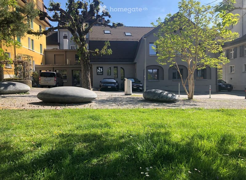 Well-maintained garden with green grass, a gravel driveway, and a modern building with a grey facade and large windows. Several cars are parked in front of the building, and there are two large, sculptural stone forms in the garden area.