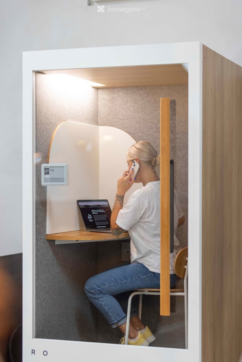 A modern, compact workspace with a laptop on a desk, a chair, and a screen displaying information. The space is enclosed with wooden and white paneling, featuring a sleek design.