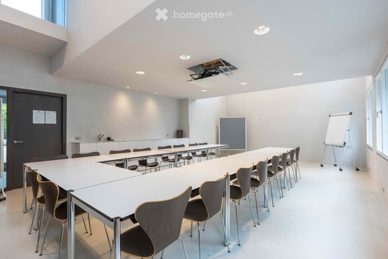 Modern conference room with white walls, large windows, and a long central table surrounded by chairs. A whiteboard and a projector are mounted on the wall, and a small sink area is visible in the background.